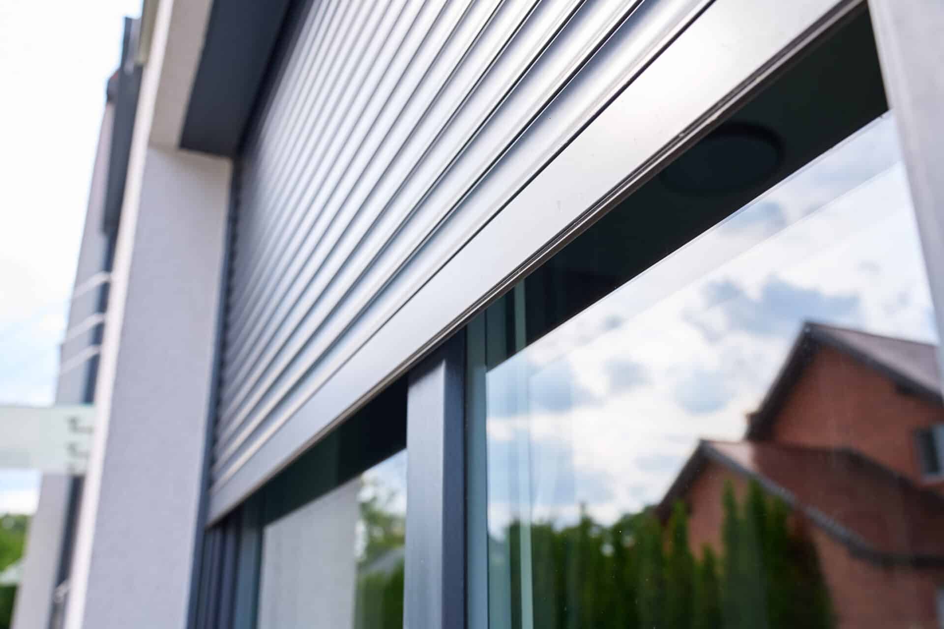A Homeowner's Guide to Privacy Glass 7 - Angelo and Sons Partially lowered metal roller shutter covers reflective exterior window showing nearby houses and cloudy sky.